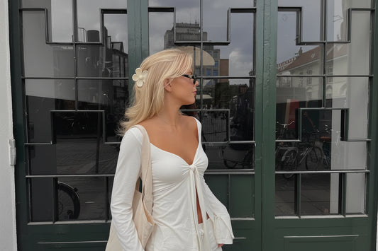 Woman in stylish white tie-front top and pants, wearing sunglasses and a flower hair clip, standing outside modern glass doors